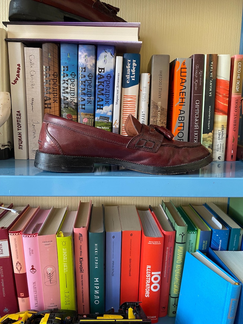 Puede incluir: Un mocas&iacute;n de cuero burdeos con detalle de borla descansa sobre una estanter&iacute;a azul, rodeado de una colecci&oacute;n de libros con lomos coloridos. El zapato es de estilo cl&aacute;sico, lo que sugiere un enfoque en calzado y accesorios.