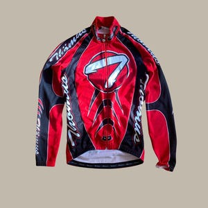 Pode incluir: Camisola de ciclismo vermelha e preta com zíper completo. A camisola apresenta um grande número sete branco na frente e o nome "Thomas" em escrita branca nas mangas. Tem uma gola alta.