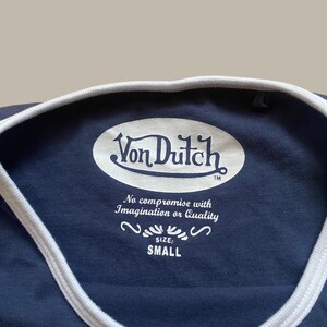 Puede incluir: Camiseta azul marino con cuello y ribete blancos. La camiseta presenta el logotipo de Von Dutch en blanco, junto con el texto "No compromise with Imagination or Quality" y la talla "SMALL".