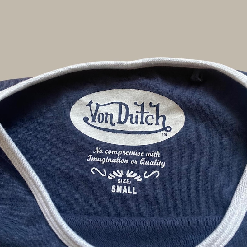 Puede incluir: Camiseta azul marino con cuello y ribete blancos. La camiseta presenta el logotipo de Von Dutch en blanco, junto con el texto "No compromise with Imagination or Quality" y la talla "SMALL".