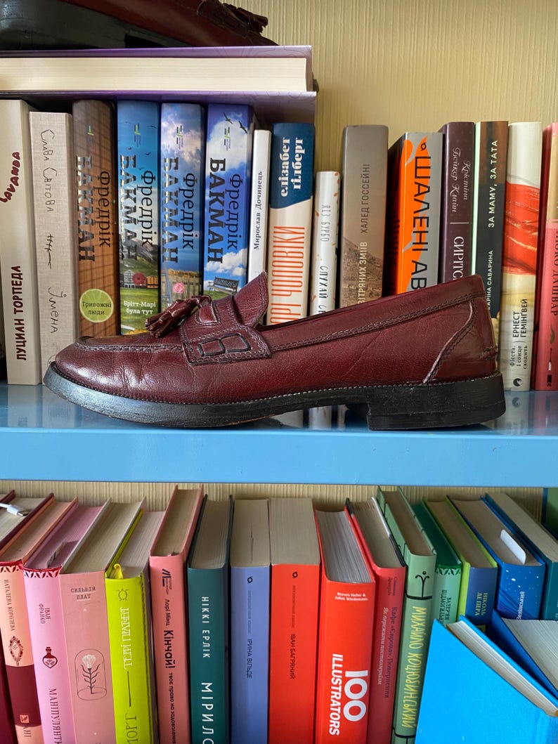 Puede incluir: Un mocas&iacute;n de cuero burdeos con detalle de borla, apoyado en una estanter&iacute;a azul frente a una estanter&iacute;a llena de libros coloridos. El zapato tiene un tac&oacute;n bajo y un dise&ntilde;o cl&aacute;sico.