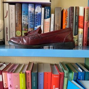 Puede incluir: Un par de mocasines de cuero burdeos con borlas, sobre una estanter&iacute;a azul. Los zapatos se exhiben frente a una colecci&oacute;n de libros coloridos. La imagen muestra un estilo cl&aacute;sico, adecuado para diversas ocasiones.