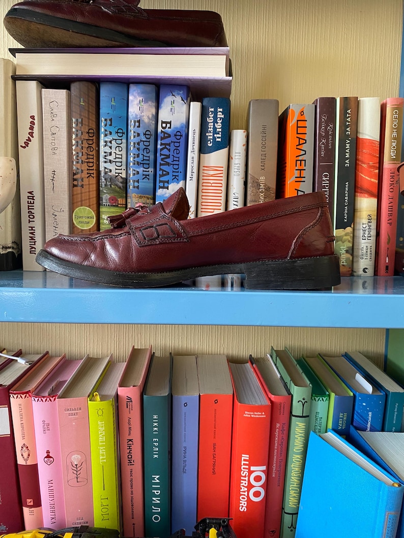 Puede incluir: Un par de mocasines de cuero burdeos con borlas, sobre una estanter&iacute;a azul. Los zapatos se exhiben frente a una colecci&oacute;n de libros coloridos. La imagen muestra un estilo cl&aacute;sico, adecuado para diversas ocasiones.