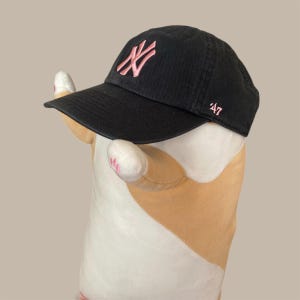 Könnte beinhalten: Schwarze Baseballkappe mit einem rosa gestickten "NY"-Logo auf der Vorderseite. Die Kappe hat einen gebogenen Schirm und die Zahl "47" auf der Seite gestickt. Die Kappe befindet sich auf einem Stofftier.