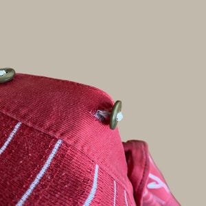 May include: Close-up of a red fabric with white vertical stripes. The fabric has a textured appearance and is secured with olive green buttons. The buttons are attached with white thread. The overall design suggests a garment or textile product.