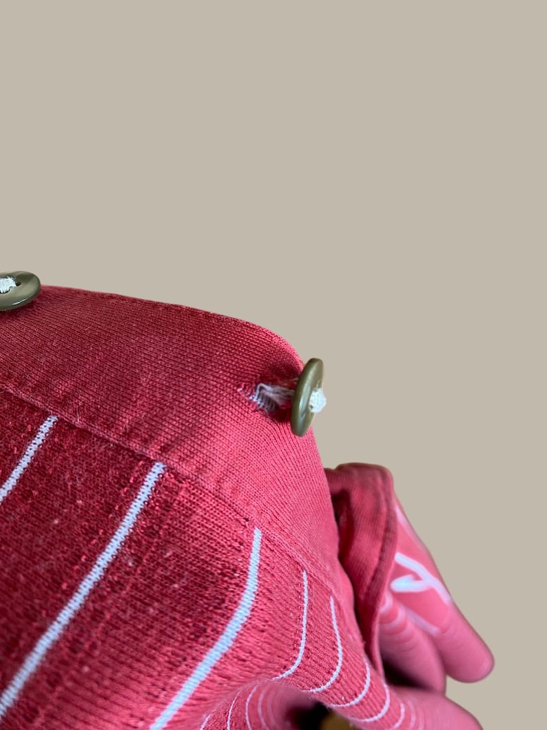 May include: Close-up of a red fabric with white vertical stripes. The fabric has a textured appearance and is secured with olive green buttons. The buttons are attached with white thread. The overall design suggests a garment or textile product.