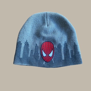 May include: Gray knit beanie featuring a red Spider-Man mask emblem with white eye details. The beanie has a cityscape design along the bottom edge. A comfortable and stylish accessory for fans of the superhero.