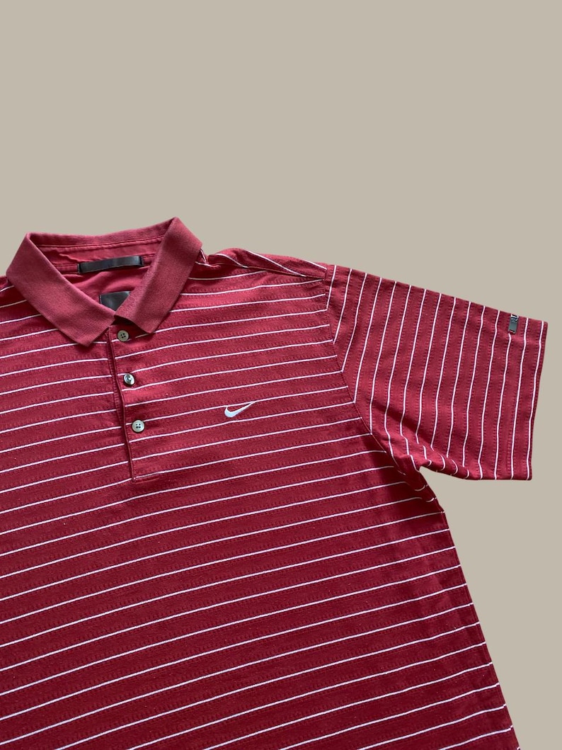 May include: A maroon polo shirt with white horizontal stripes. The shirt has a collar, three buttons, and short sleeves. A small white Nike swoosh logo is visible on the chest.