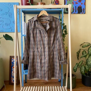 Puede incluir: Un abrigo tipo trench a cuadros en tonos marrón, rojo y azul cuelga de una percha de madera. El abrigo presenta un diseño cruzado con botones grandes y un cuello. El abrigo se exhibe en un perchero de madera.