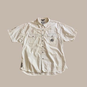 Op de afbeelding: Crèmekleurig overhemd met korte mouwen, twee borstzakken en kleine geborduurde patches. Het overhemd heeft een klassieke kraag en een relaxte pasvorm, geschikt voor casual wear.