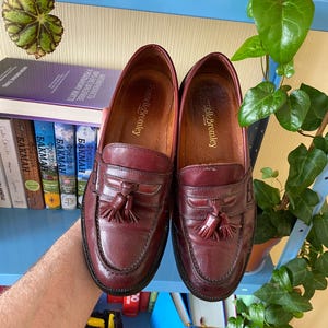 Puede incluir: Un par de mocasines de cuero burdeos con detalles de borlas. Los zapatos tienen un dise&ntilde;o cl&aacute;sico con punta redonda y tac&oacute;n bajo. La marca "Russell & Bromley" es visible. Los zapatos son sostenidos por una persona.
