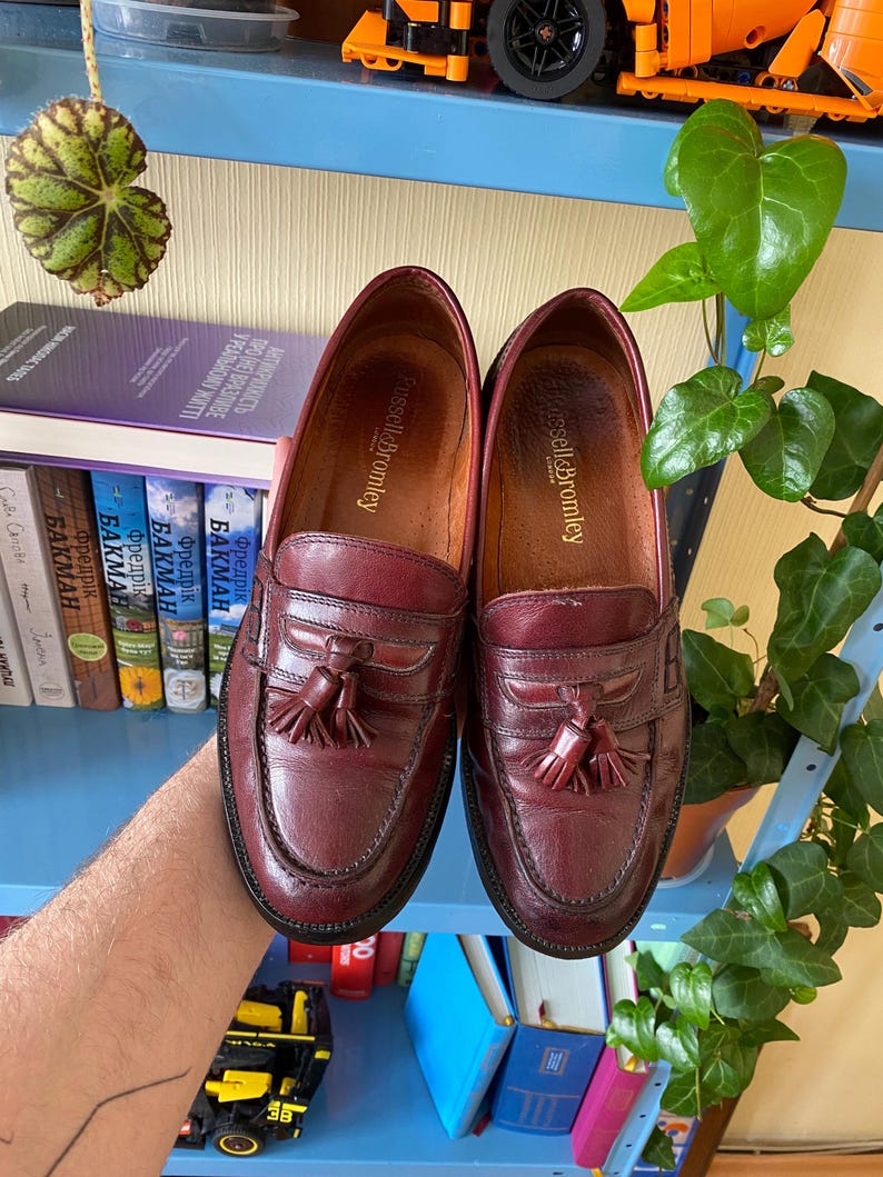 Puede incluir: Un par de mocasines de cuero burdeos con detalles de borlas. Los zapatos tienen un dise&ntilde;o cl&aacute;sico con punta redonda y tac&oacute;n bajo. La marca "Russell & Bromley" es visible. Los zapatos son sostenidos por una persona.