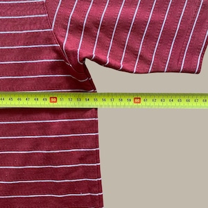 May include: A maroon and white striped short-sleeve shirt. The shirt has horizontal stripes on the lower portion and vertical stripes on the upper portion. A yellow measuring tape is placed across the bottom of the shirt.