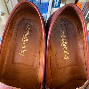 Puede incluir: Primer plano de dos mocasines de cuero burdeos. El interior de cada zapato es de un marr&oacute;n m&aacute;s claro con el nombre de la marca "Russell & Bromley London" en letras doradas. Los zapatos parecen desgastados.