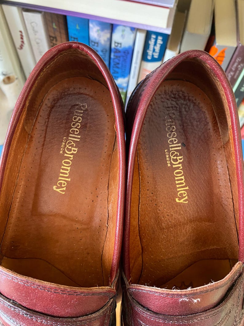 Puede incluir: Primer plano de dos mocasines de cuero burdeos. El interior de cada zapato es de un marr&oacute;n m&aacute;s claro con el nombre de la marca "Russell & Bromley London" en letras doradas. Los zapatos parecen desgastados.