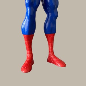 Puede incluir: Primer plano de la parte inferior del cuerpo de una figura de acci&oacute;n de Spider-Man. Las piernas de la figura son azules, que pasan a pies y pantorrillas rojas con un patr&oacute;n de telara&ntilde;a. La figura est&aacute; sobre un fondo neutro.