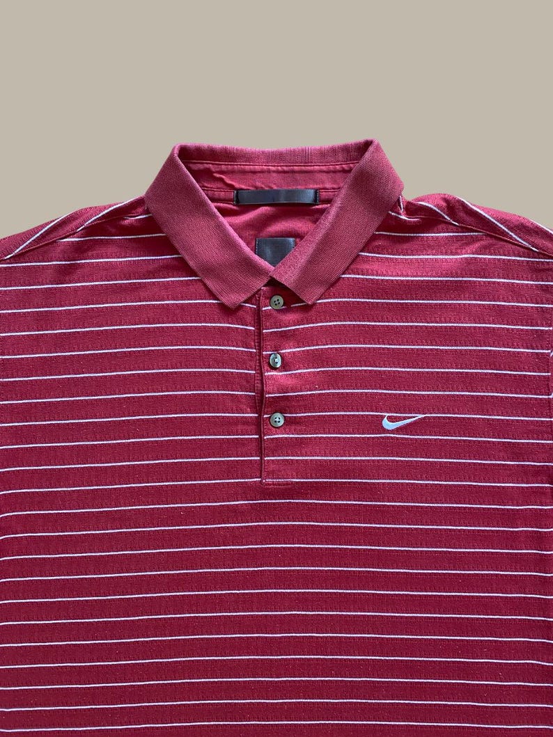May include: Maroon polo shirt with white horizontal stripes. Features a collared neckline, three-button placket, and a small Nike logo on the chest. The shirt has a classic, sporty design.