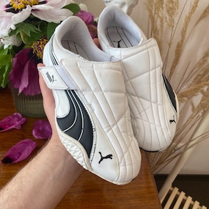 Könnte beinhalten: Weiße und schwarze Puma-Sneaker mit Steppdesign. Die Sneaker haben einen Klettverschluss und das Puma-Logo. Die Schuhe werden auf einer Holzoberfläche gezeigt.