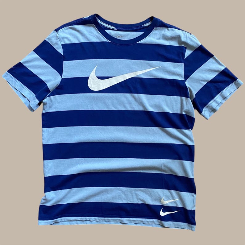 K&ouml;nnte beinhalten: Blau-hellblau gestreiftes T-Shirt mit wei&szlig;em Nike Swoosh-Logo auf der Brust. Das Shirt hat kurze &Auml;rmel und einen Rundhalsausschnitt. Ein kleineres Nike Swoosh-Logo ist unten sichtbar.