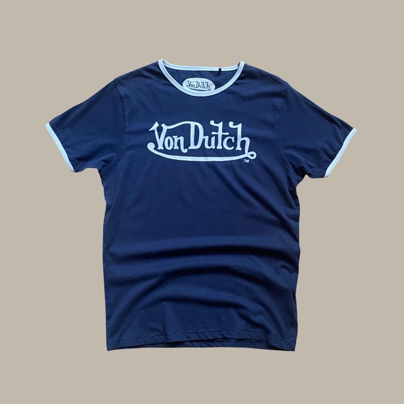 Puede incluir: Camiseta azul marino con ribetes blancos en el cuello y las mangas. La camiseta presenta el texto blanco "Von Dutch" en una escritura estilizada en la parte delantera. Un logotipo m&aacute;s peque&ntilde;o de "Von Dutch" est&aacute; en el cuello.