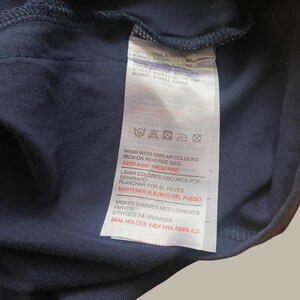 Puede incluir: Una etiqueta de prenda azul marino con instrucciones de cuidado en varios idiomas. La etiqueta incluye s&iacute;mbolos de lavado, planchado y seguridad contra incendios. El texto incluye "100% COTTON" y "KEEP AWAY FROM FIRE."