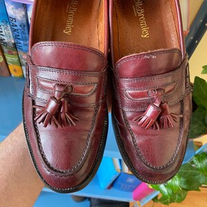 Puede incluir: Un par de mocasines de cuero burdeos con detalles de borlas. Los zapatos tienen un dise&ntilde;o cl&aacute;sico con punta redonda y suela cosida. La marca "Russell & Bromley" es visible dentro del zapato.
