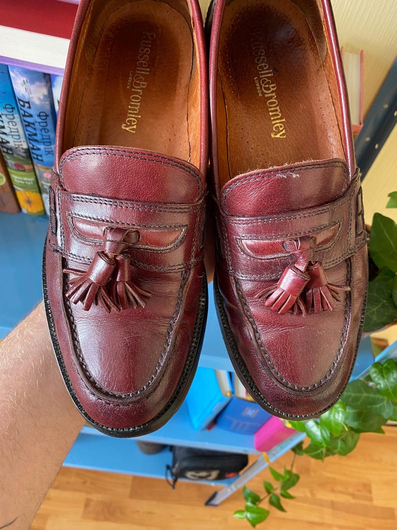Puede incluir: Un par de mocasines de cuero burdeos con detalles de borlas. Los zapatos tienen un dise&ntilde;o cl&aacute;sico con punta redonda y suela cosida. La marca "Russell & Bromley" es visible dentro del zapato.