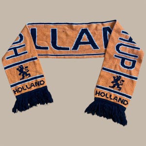May include: Orange and navy blue knitted scarf with the word "HOLLAND" and the word "UP" on the ends. The word "HOLLAND" is also written on the bottom of each end of the scarf. The scarf has a navy blue fringe.