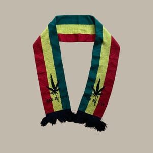 May include: A knitted scarf in the colors of red, yellow, and green, with black fringe. The scarf features a cannabis leaf design and the words "Peace and Love" in black.