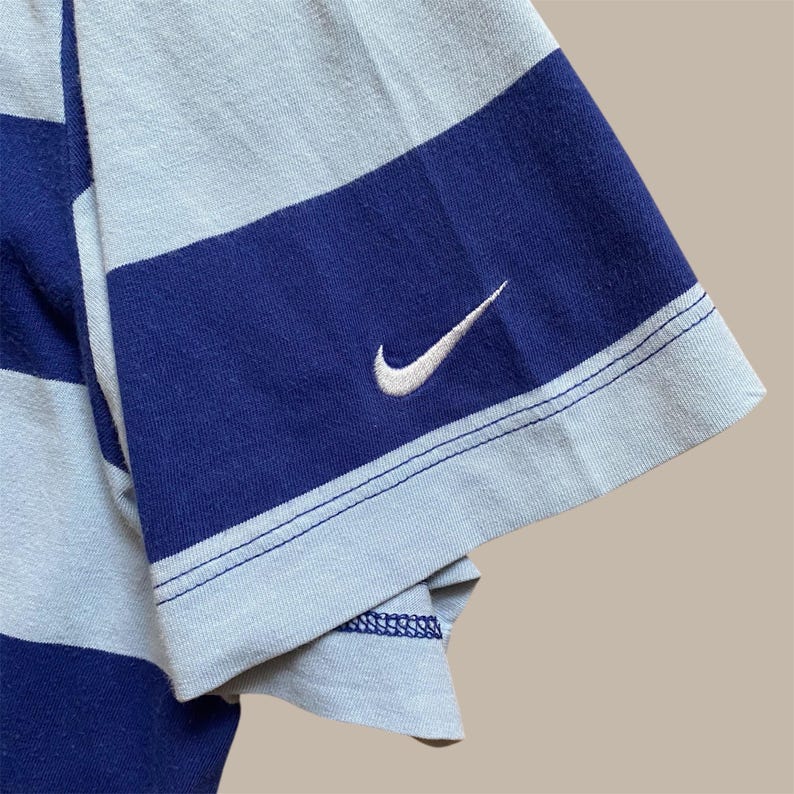 K&ouml;nnte beinhalten: Nahaufnahme eines blau-wei&szlig; gestreiften T-Shirt-&Auml;rmels. Der &Auml;rmel hat ein wei&szlig;es Nike Swoosh-Logo, das auf den blauen Streifen gestickt ist. Das Shirt hat ein klassisches, sportliches Design.