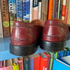 Puede incluir: Un par de mocasines de cuero burdeos con suelas negras, frente a una estanter&iacute;a de libros coloridos. Los zapatos, con sus costuras visibles, presentan un dise&ntilde;o cl&aacute;sico.