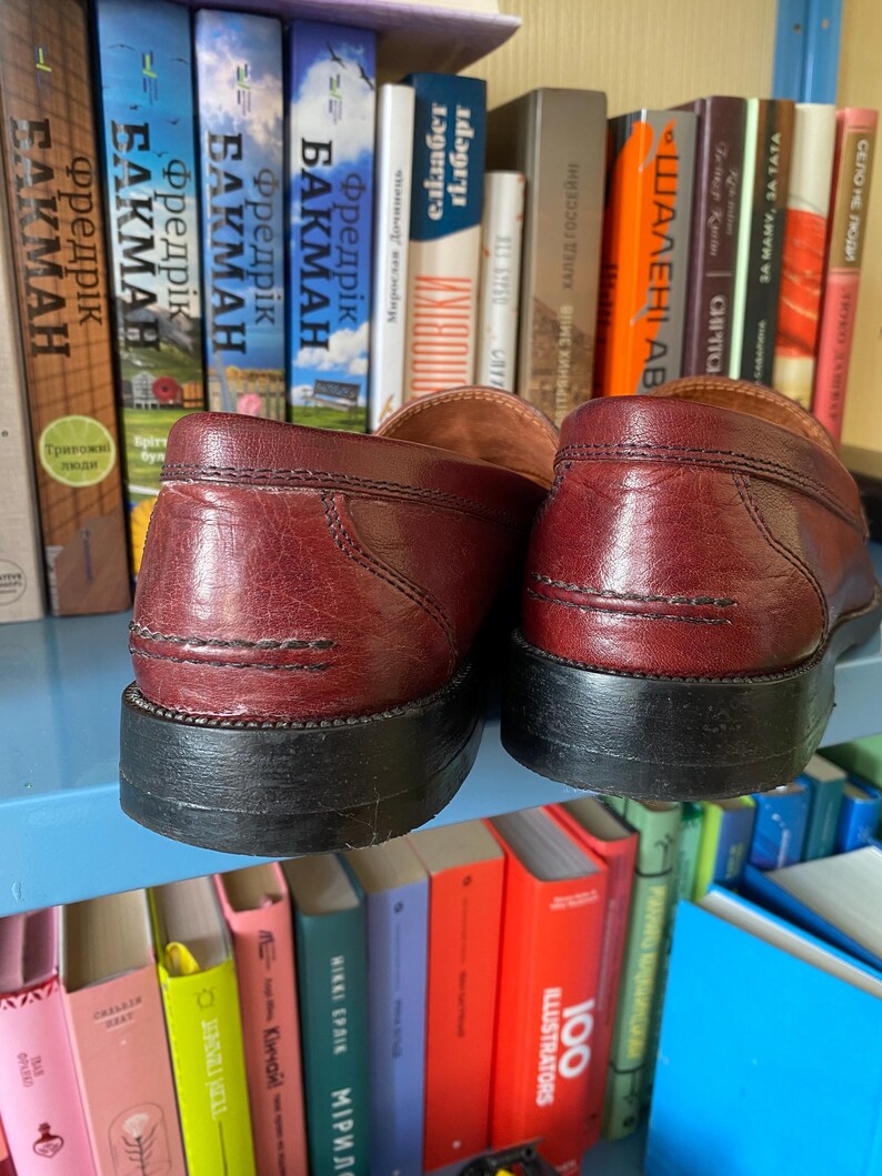 Puede incluir: Un par de mocasines de cuero burdeos con suelas negras, frente a una estanter&iacute;a de libros coloridos. Los zapatos, con sus costuras visibles, presentan un dise&ntilde;o cl&aacute;sico.