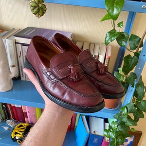 Puede incluir: Un par de mocasines de cuero burdeos con suelas negras. Cada zapato presenta una borla decorativa y una correa de mocas&iacute;n penny. Los zapatos est&aacute;n en la mano de una persona.
