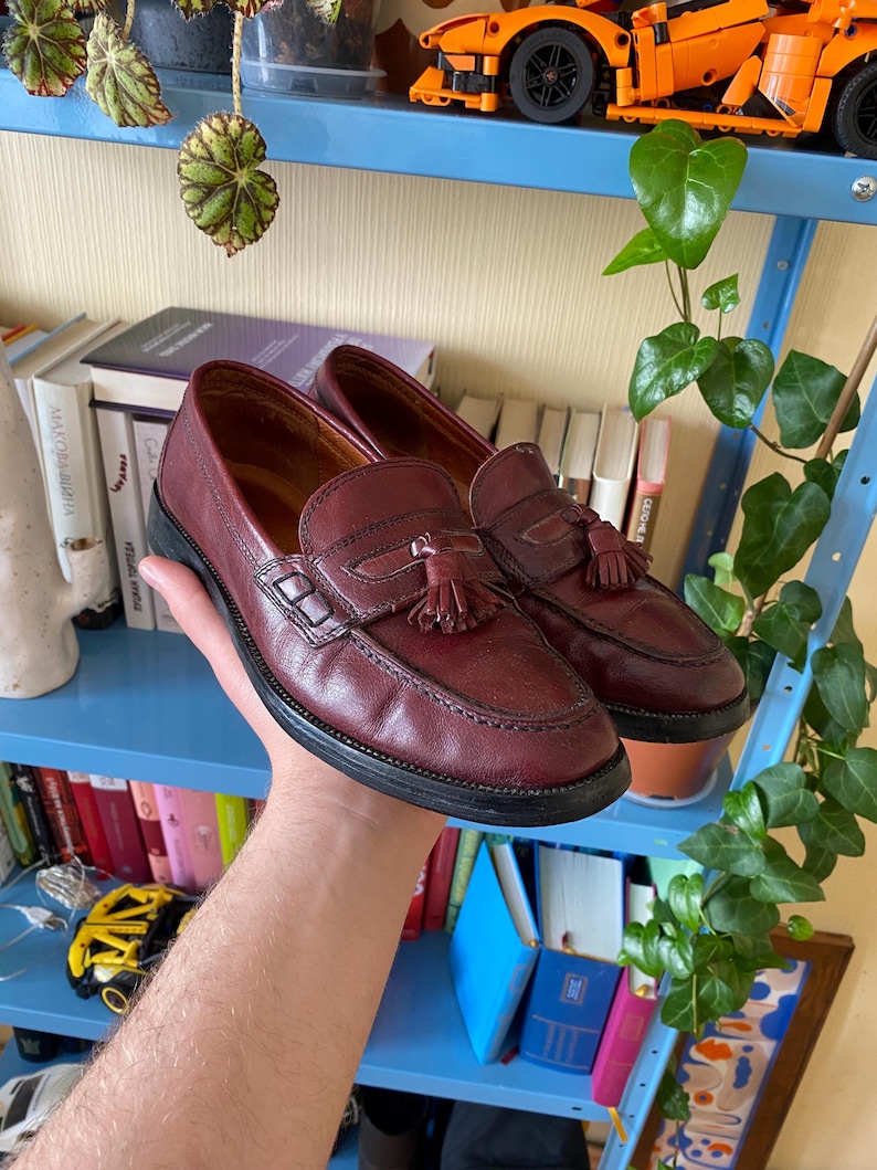 Puede incluir: Un par de mocasines de cuero burdeos con suelas negras. Cada zapato presenta una borla decorativa y una correa de mocas&iacute;n penny. Los zapatos est&aacute;n en la mano de una persona.