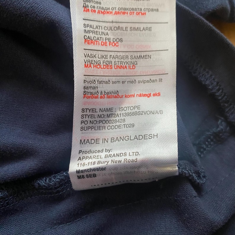 Puede incluir: Una etiqueta de prenda azul marino con texto en varios idiomas. La etiqueta incluye instrucciones de cuidado, el nombre del estilo "ISOTOPE", el n&uacute;mero de estilo, el n&uacute;mero de pedido, el c&oacute;digo del proveedor y "MADE IN BANGLADESH".