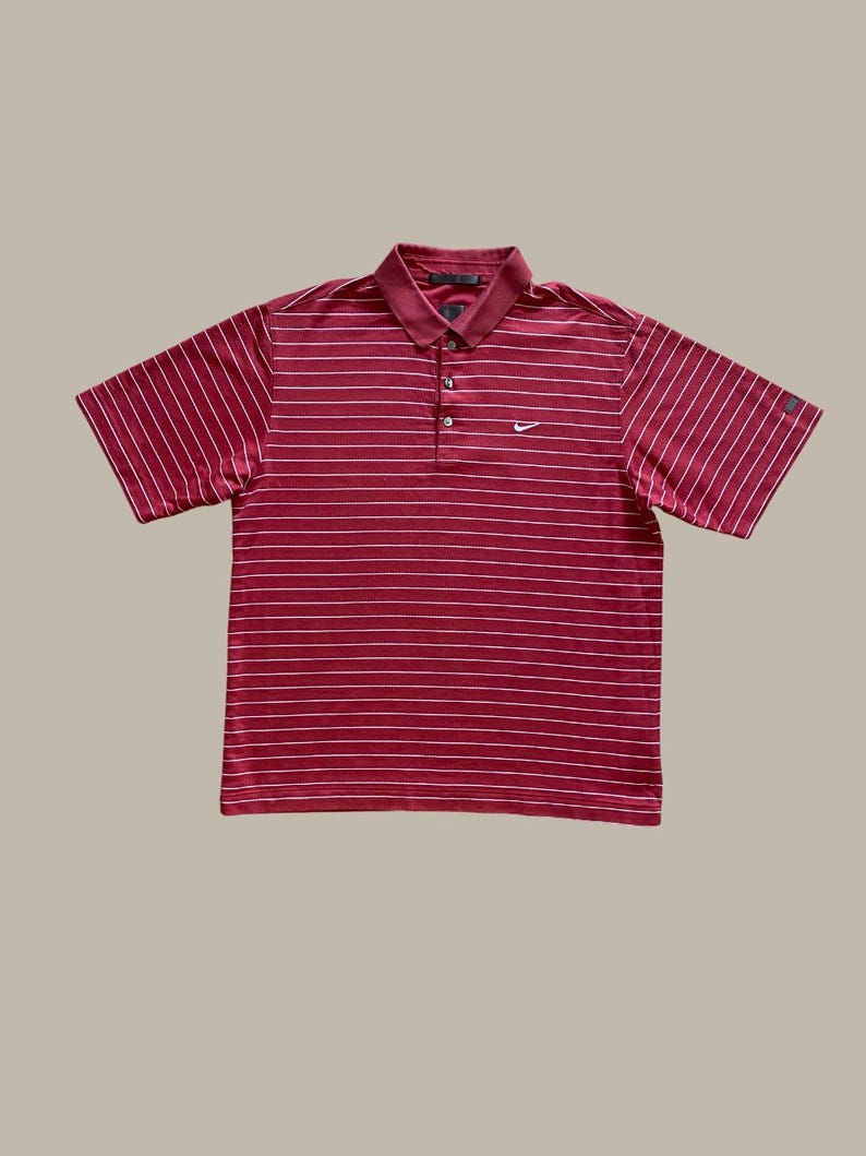 May include: A short-sleeved polo shirt in a deep red hue, featuring horizontal white stripes. The shirt has a collared neckline, a three-button placket, and a small Nike logo on the chest. The overall design is classic and sporty.