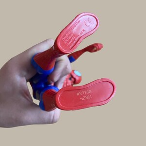 Puede incluir: Una figura de acci&oacute;n de Spider-Man roja, azul y negra. Se ven los pies de la figura, con el texto "#B9760 62861" y "TM&&copy; 2013 Marvel & Subs" impreso en las suelas. La figura se sostiene en una mano.