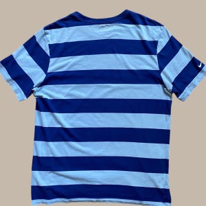 K&ouml;nnte beinhalten: Blau-wei&szlig; gestreiftes T-Shirt mit kurzen &Auml;rmeln. Das Shirt hat einen Rundhalsausschnitt und ein kleines wei&szlig;es Nike-Logo auf dem &Auml;rmel. Die Streifen sind horizontal und gleichm&auml;&szlig;ig angeordnet.