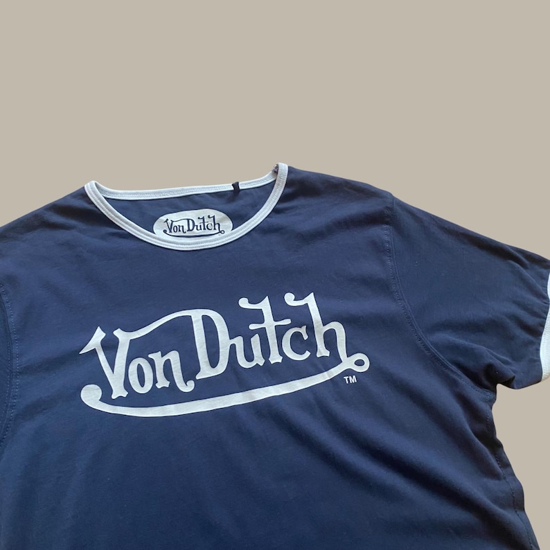 Puede incluir: Camiseta azul marino con ribete blanco y la inscripci&oacute;n "Von Dutch" en blanco, con una tipograf&iacute;a estilizada. La camiseta tiene cuello redondo y un peque&ntilde;o logotipo "Von Dutch" cerca del cuello.