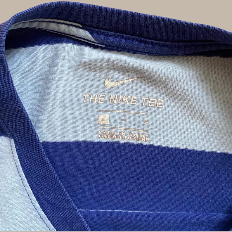 K&ouml;nnte beinhalten: Nahaufnahme eines blau-wei&szlig; gestreiften T-Shirts mit dem Nike-Logo und dem Aufdruck "THE NIKE TEE" auf der Vorderseite. Das Shirt hat einen Rundhalsausschnitt und ein Etikett mit der Aufschrift "MADE IN CHINA".