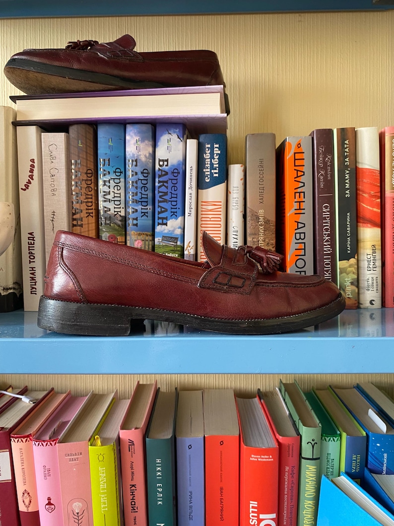 Puede incluir: Un par de mocasines de cuero burdeos con detalles de borlas, exhibidos en una estanter&iacute;a azul sobre una colecci&oacute;n de libros. Los zapatos son de estilo cl&aacute;sico, mostrando un color rico y un dise&ntilde;o tradicional.
