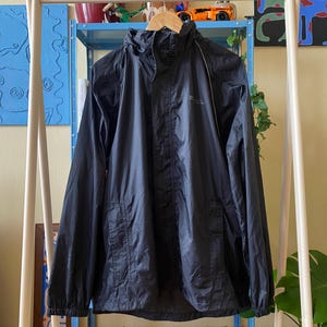 Może przedstawiać: Czarna kurtka przeciwdeszczowa z kapturem, zapięciem na zamek i długimi rękawami. Kurtka wisi na drewnianym wieszaku. Kurtka ma małe logo na piersi. Kurtka jest wykonana z lekkiego materiału.