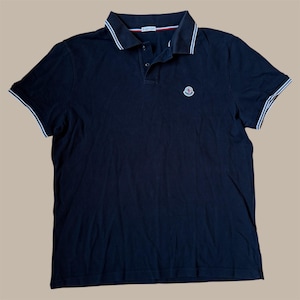 Könnte beinhalten: Marineblaues Poloshirt mit weißem Besatz an Kragen und Ärmeln. Das Shirt hat eine Knopfleiste mit drei Knöpfen und ein kleines Logo auf der Brust. Der Kragen hat einen rot-weiß-blauen Streifen.