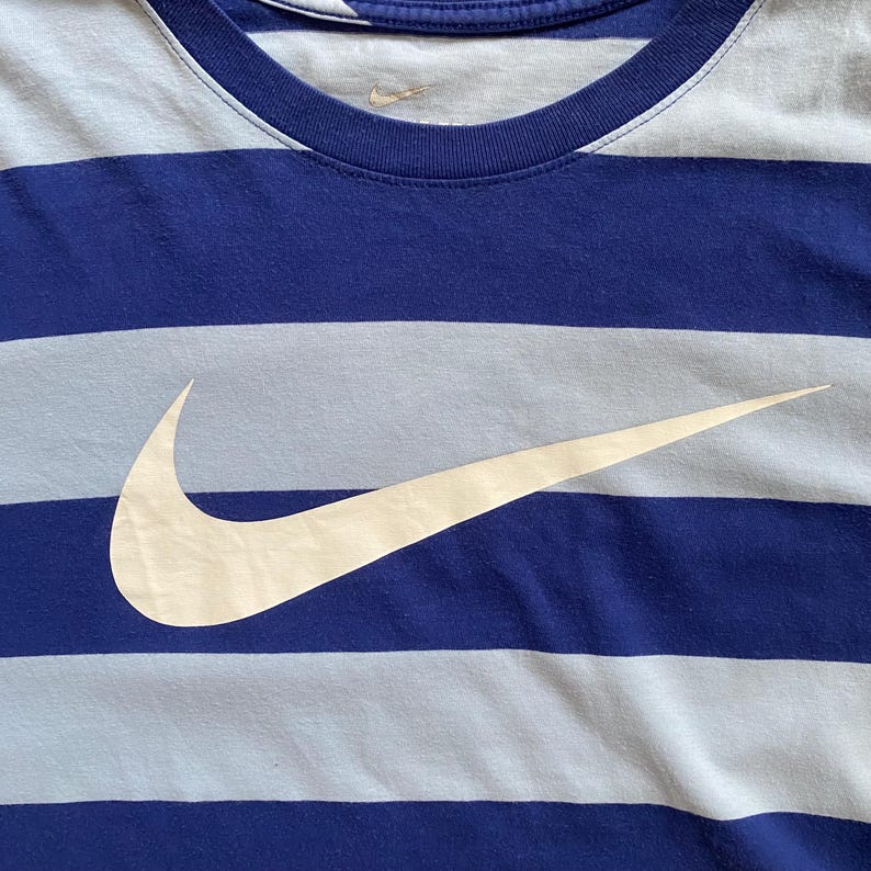 K&ouml;nnte beinhalten: Blau-wei&szlig; gestreiftes T-Shirt mit wei&szlig;em Nike-Logo. Das Shirt hat einen Rundhalsausschnitt und kurze &Auml;rmel. Der Nike-Swoosh ist prominent auf der Vorderseite des Shirts zu sehen.