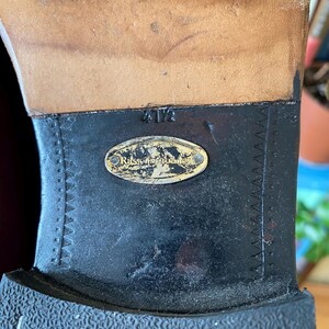 Puede incluir: Primer plano de una suela de zapato, con una parte superior de cuero desgastada y una suela de goma negra. La suela tiene un patr&oacute;n texturizado y una etiqueta met&aacute;lica con el nombre de la marca. La talla es 4 1/2.