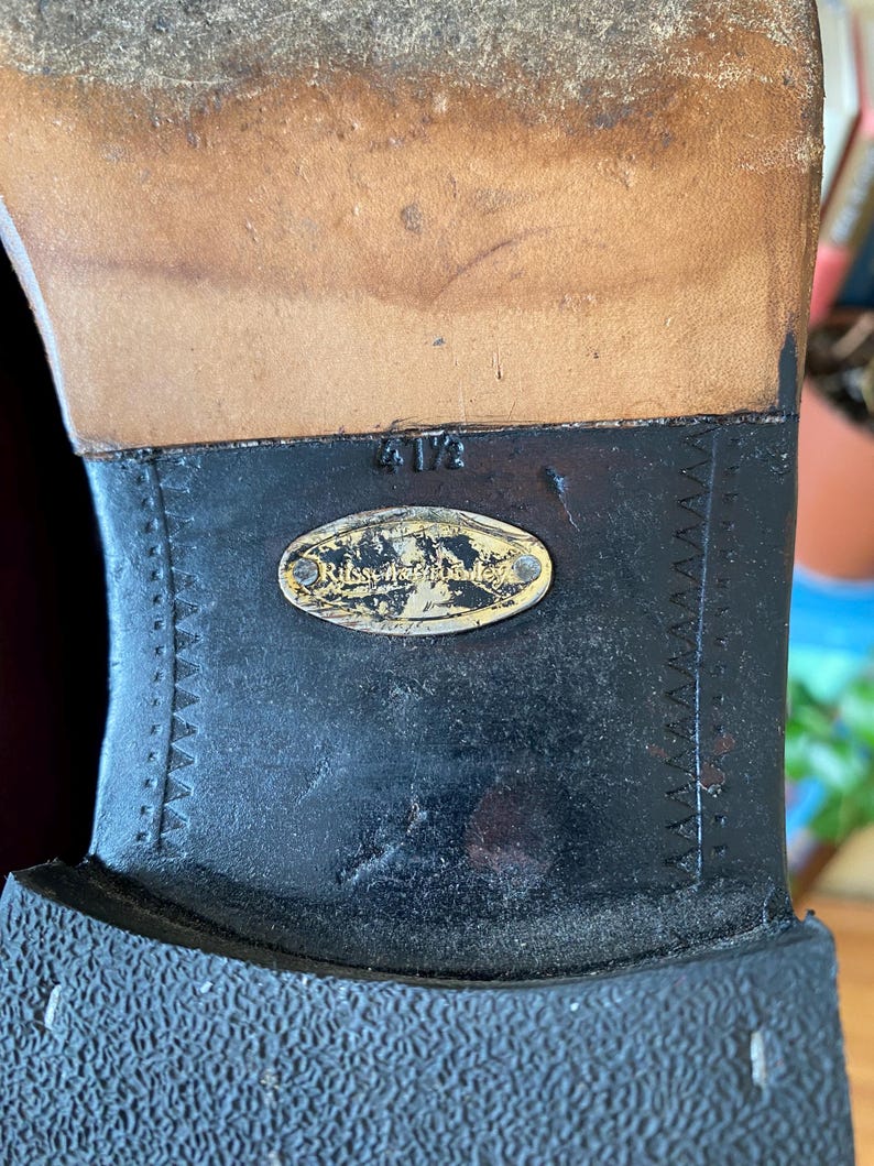 Puede incluir: Primer plano de una suela de zapato, con una parte superior de cuero desgastada y una suela de goma negra. La suela tiene un patr&oacute;n texturizado y una etiqueta met&aacute;lica con el nombre de la marca. La talla es 4 1/2.