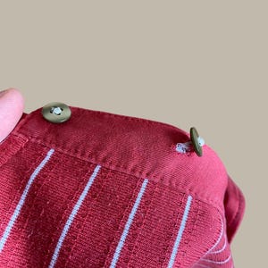 May include: Close-up of a red collared shirt with vertical white stripes. The shirt has two olive green buttons on the collar. The fabric has a textured appearance.