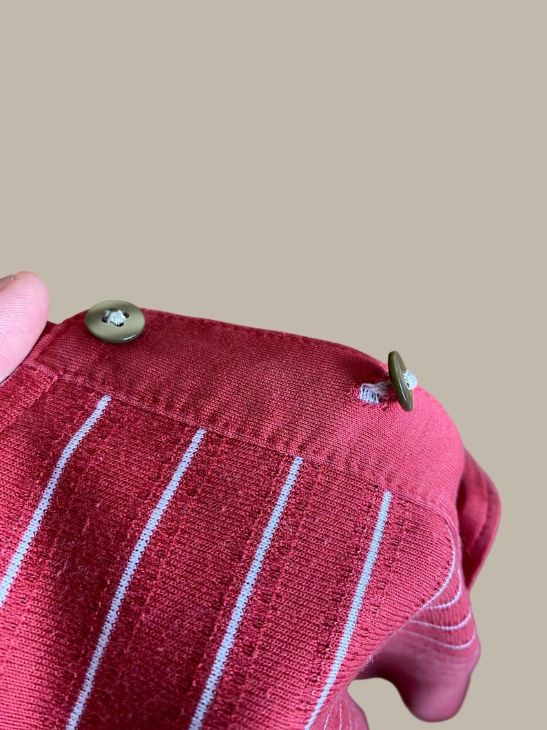 May include: Close-up of a red collared shirt with vertical white stripes. The shirt has two olive green buttons on the collar. The fabric has a textured appearance.