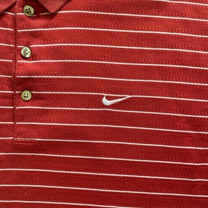 May include: Red polo shirt with white horizontal stripes. Features a three-button placket and a white Nike swoosh logo on the chest. The shirt has a ribbed collar and is made of a textured fabric.