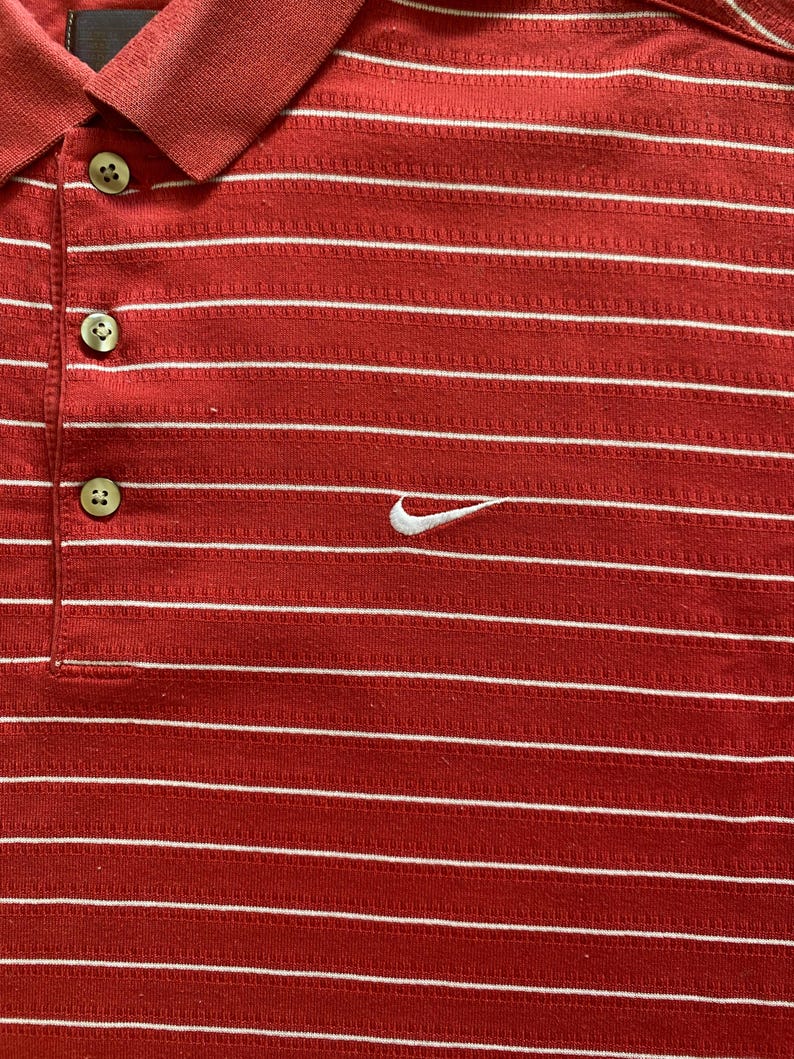May include: Red polo shirt with white horizontal stripes. Features a three-button placket and a white Nike swoosh logo on the chest. The shirt has a ribbed collar and is made of a textured fabric.